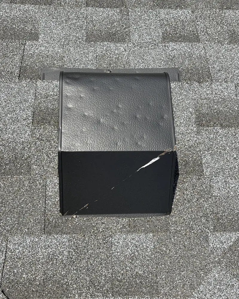 Roof vent with hail dents and a cracked seam on a gray shingle roof